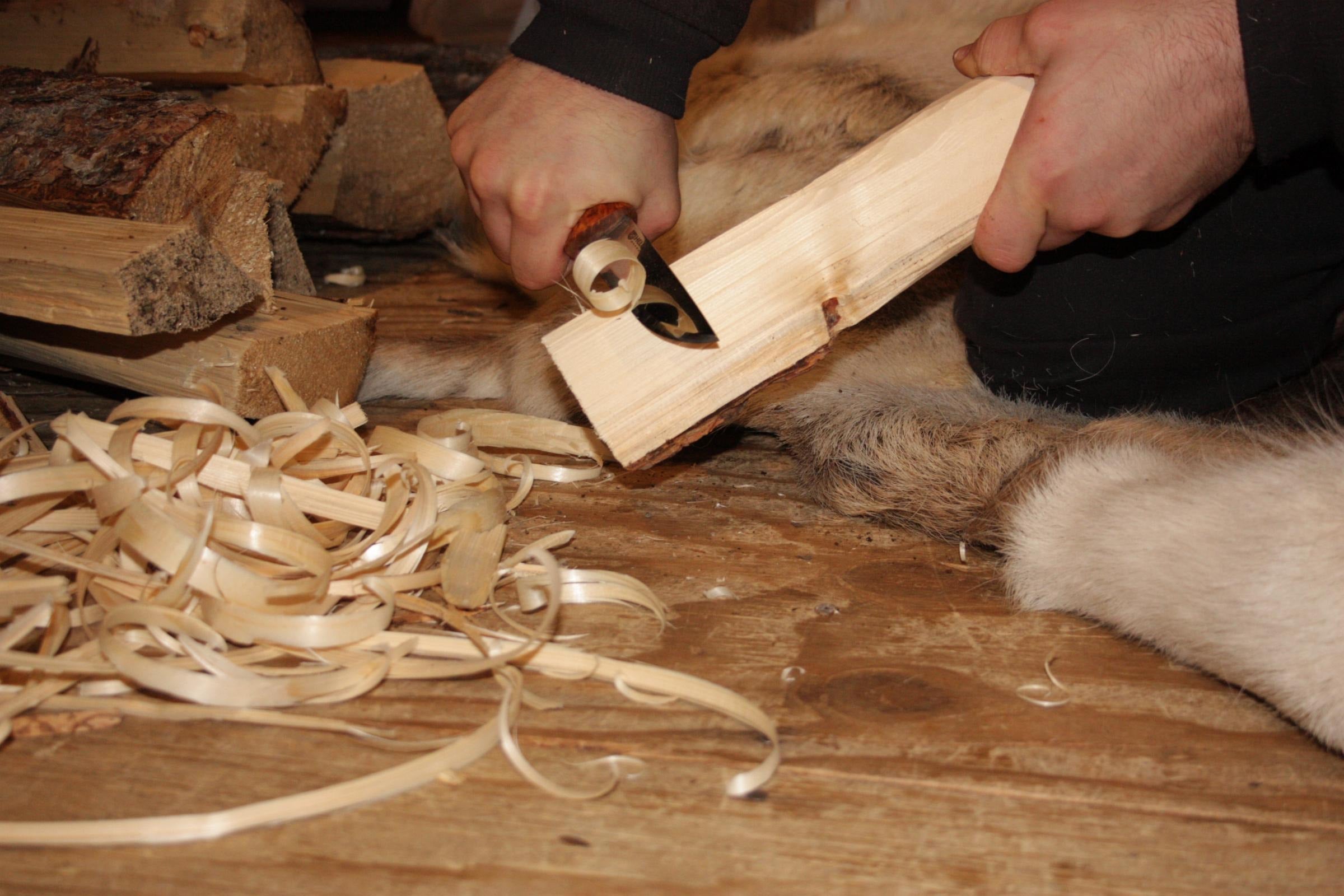 Person carving wood with knife on fur rug, outdoor survival gear