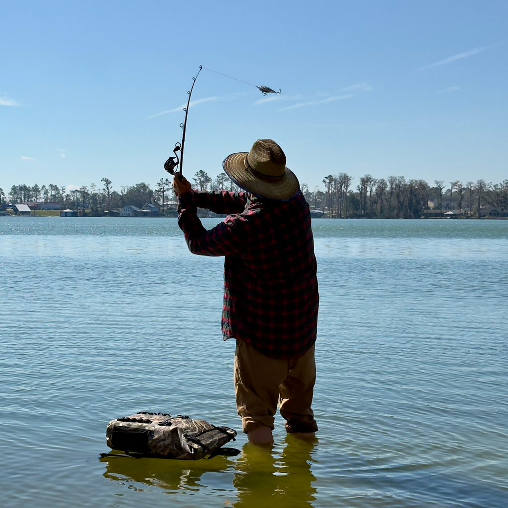Angler in red plaid shirt and straw hat casting fishing rod from shallow water, camo tackle bag nearby