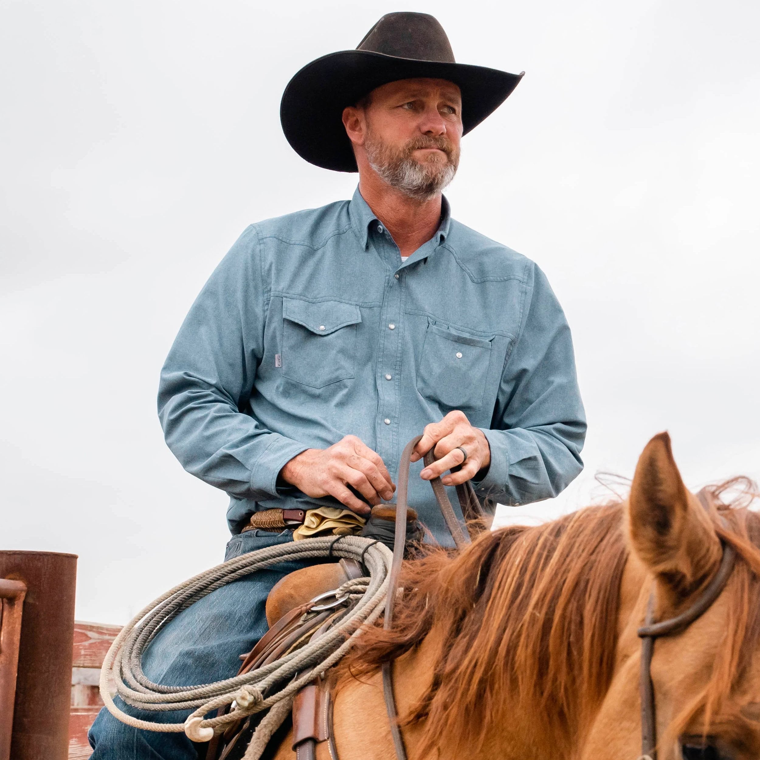 Man in blue long sleeve shirt and cowboy hat riding horse with lasso, outdoor gear for hunting and ranch use