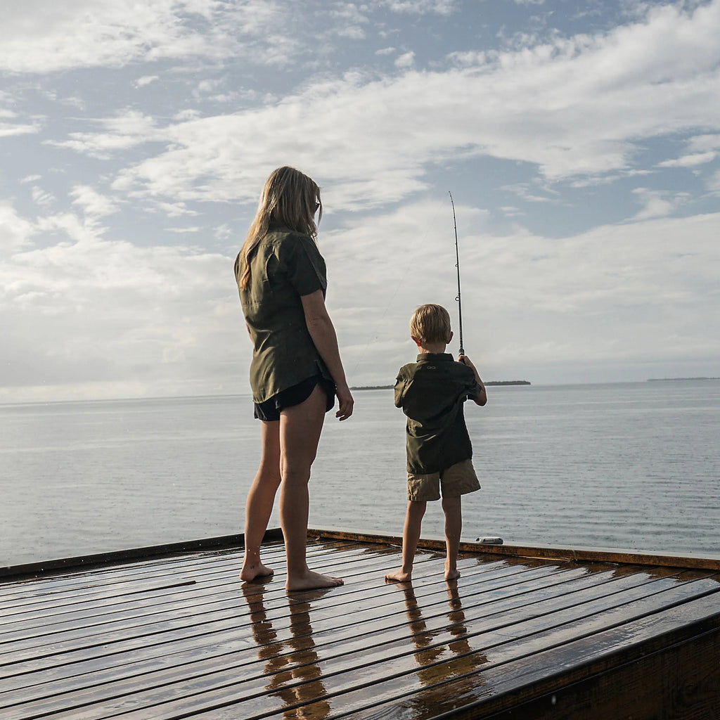 Woman and boy in olive shirts fishing from wooden dock on calm water under cloudy sky