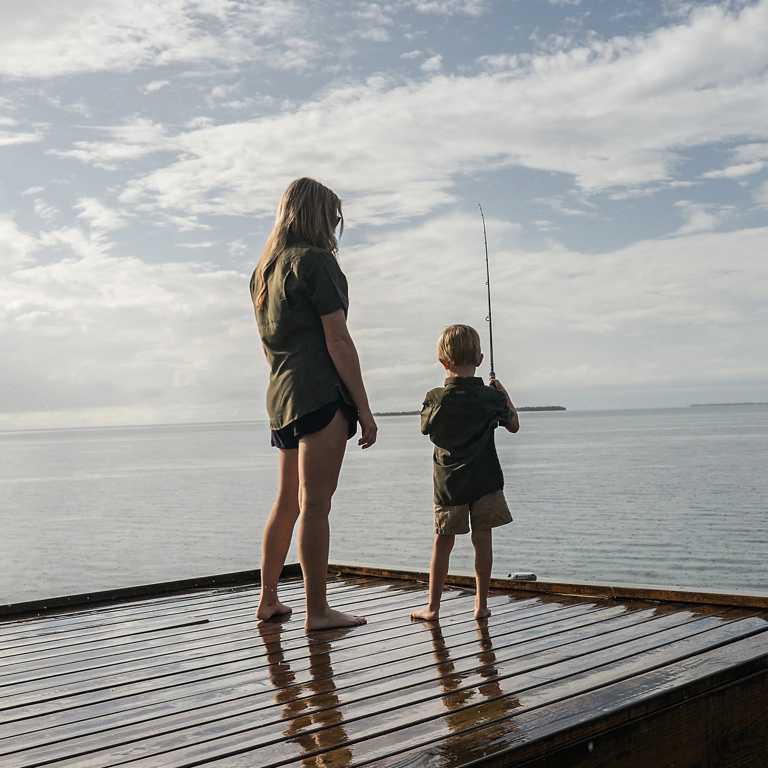 Woman and boy in olive shirts fishing from wooden dock on calm water under cloudy sky
