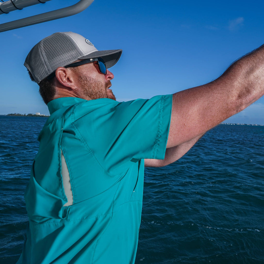 Man wearing teal fishing shirt and gray trucker hat on boat, casting line under blue sky