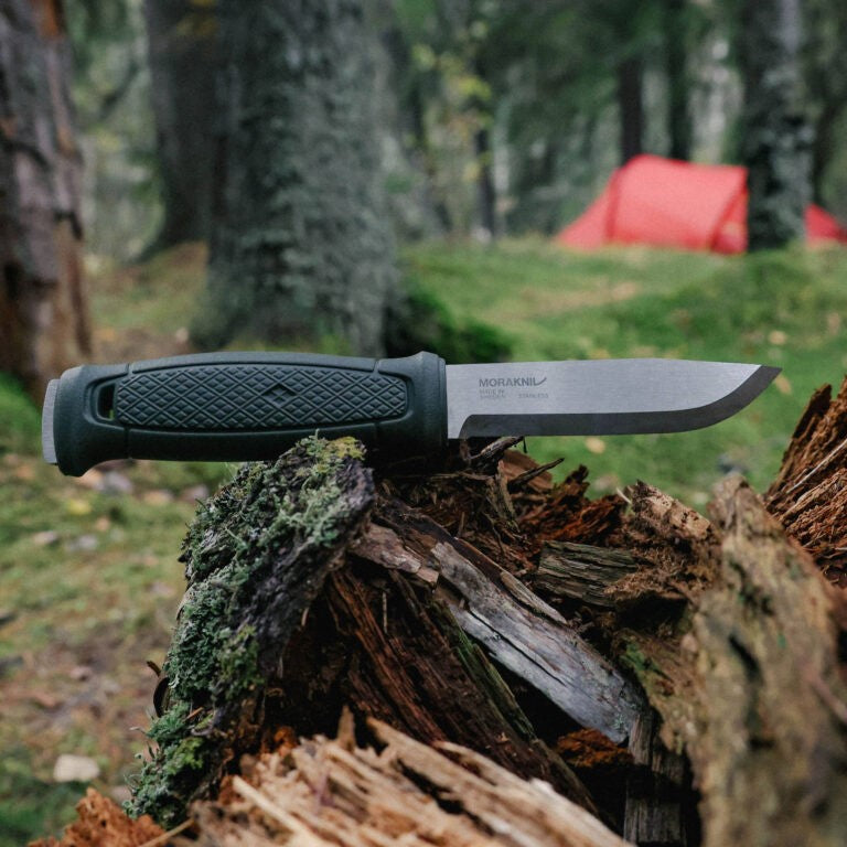 Morakniv outdoor knife with black grip, stainless steel blade, resting on mossy log in forest camp setting