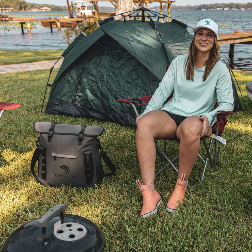 Woman in mint long sleeve shirt, coral HUK boots, white cap, sitting by green tent near lake with Dry Pocket bag
