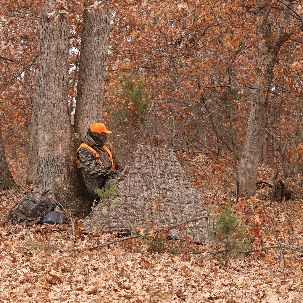 Hunter in camo gear and orange vest using Nukem ground blind in autumn woods for deer hunting