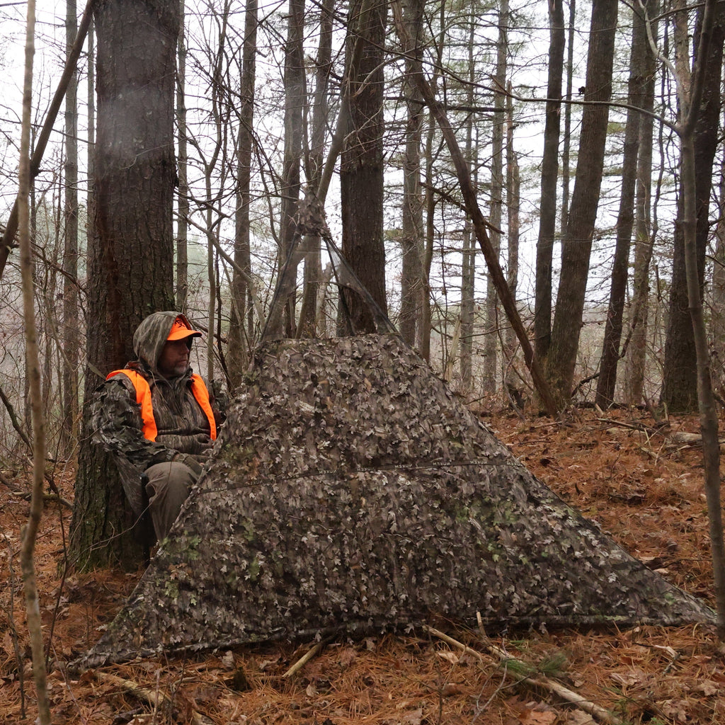 Hunting blind with leaf camo pattern in forest, hunter wearing orange safety vest and camo gear