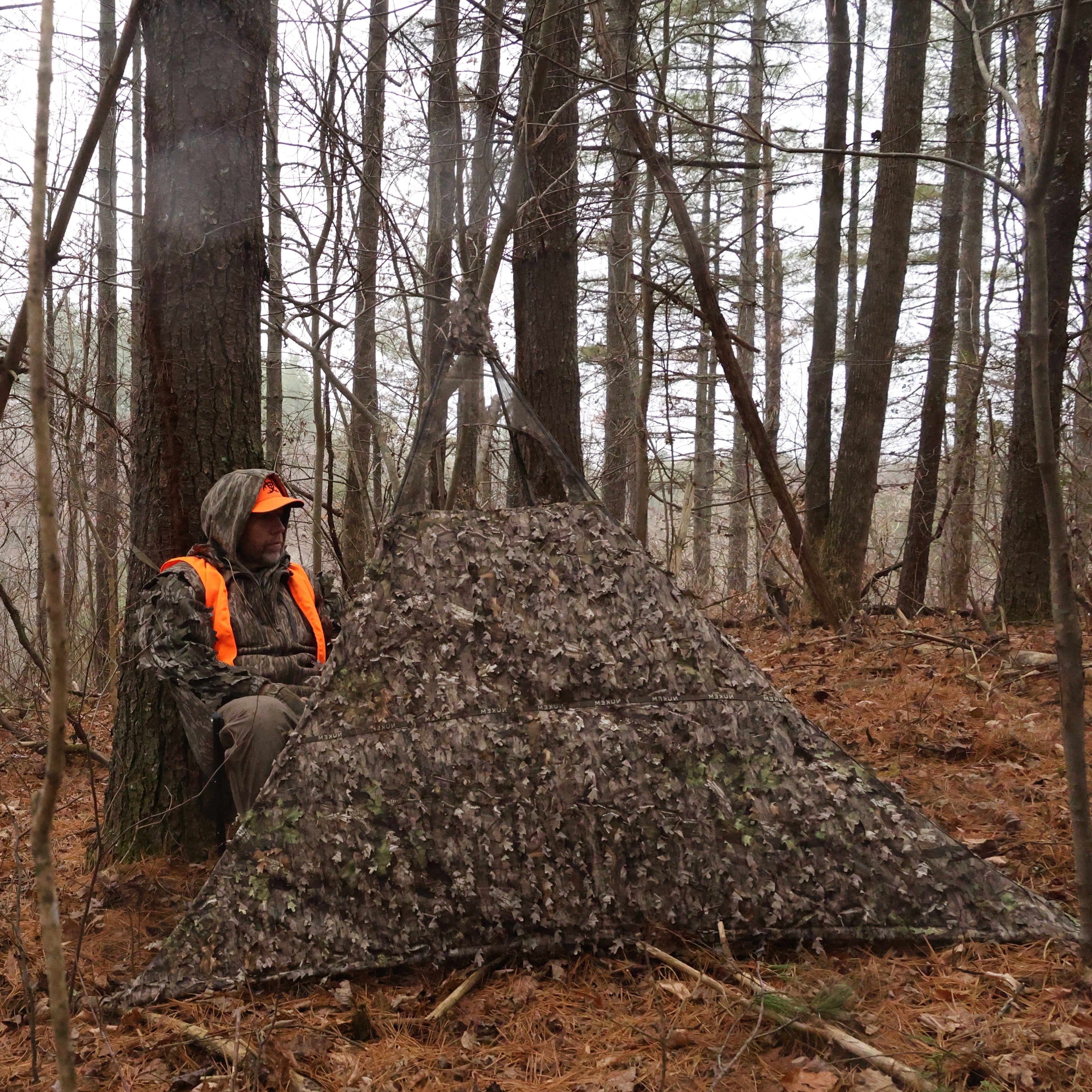 Hunting blind with leaf camo pattern in forest, hunter wearing orange safety vest and camo gear