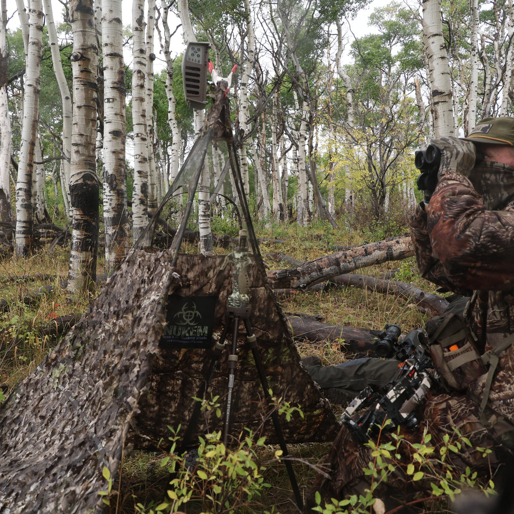 Hunter in camo using binoculars beside Nukem hunting blind in aspen forest with trail camera