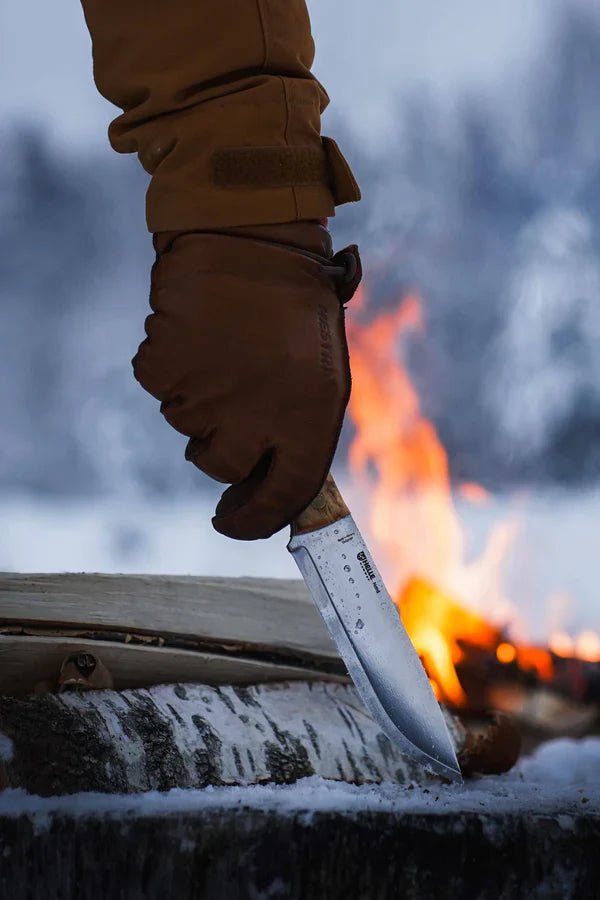Hunting knife with wooden handle, held in gloved hand near campfire in snowy woods