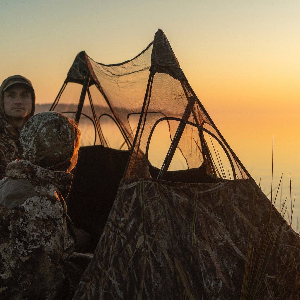 Camo hunting blind with mesh windows for waterfowl hunting at sunrise, worn by hunters in marsh gear