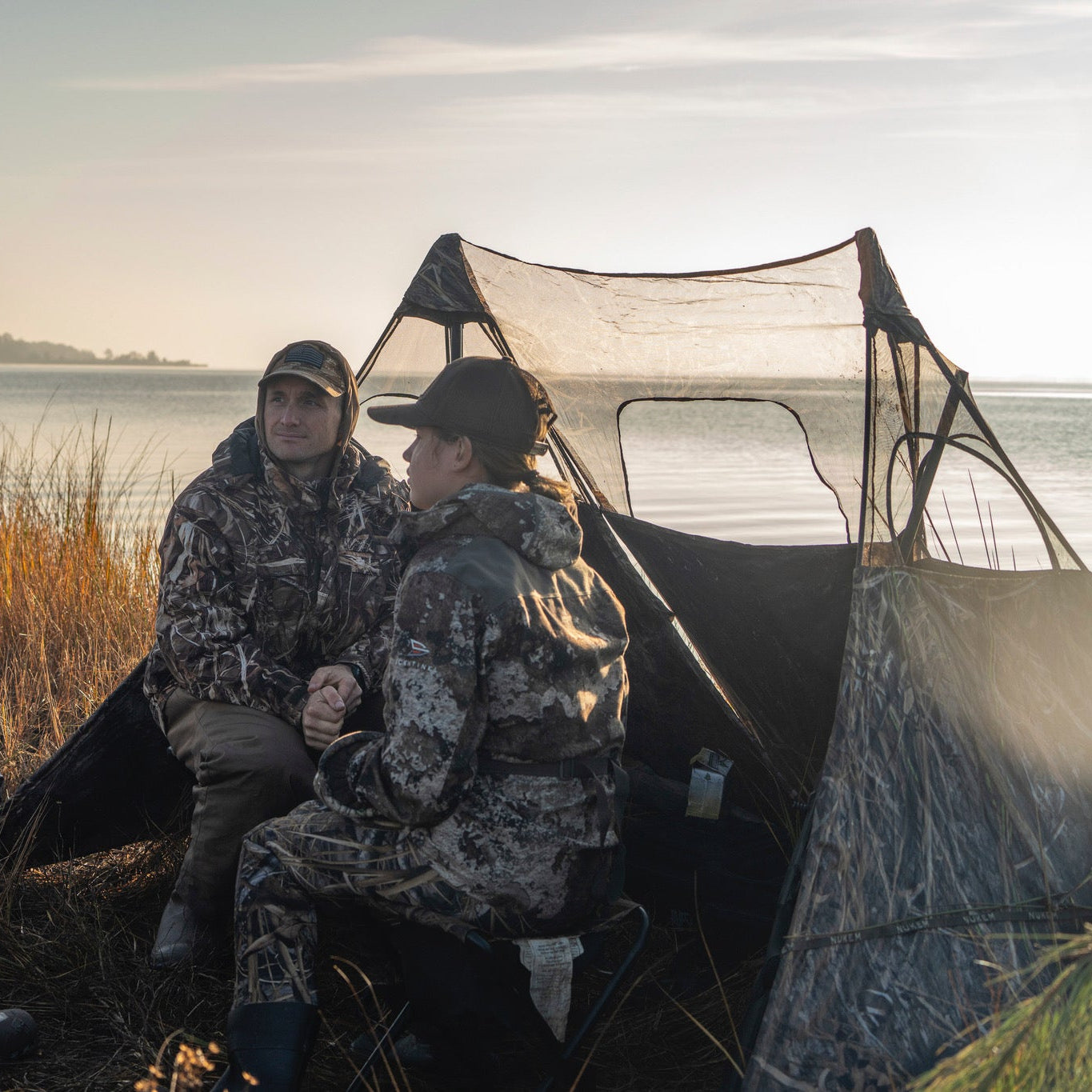 Hunters in camo gear sit by water near a camo hunting blind at sunrise