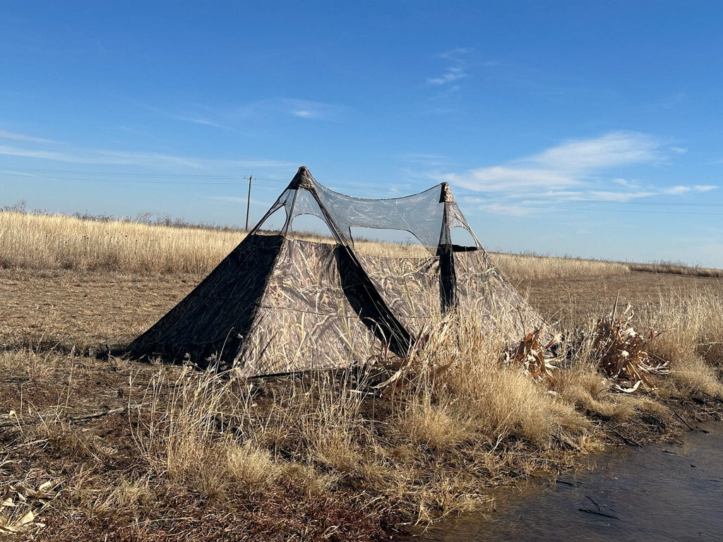 Camo hunting blind tent with mesh windows set up in dry field near water, ideal for waterfowl or deer hunting