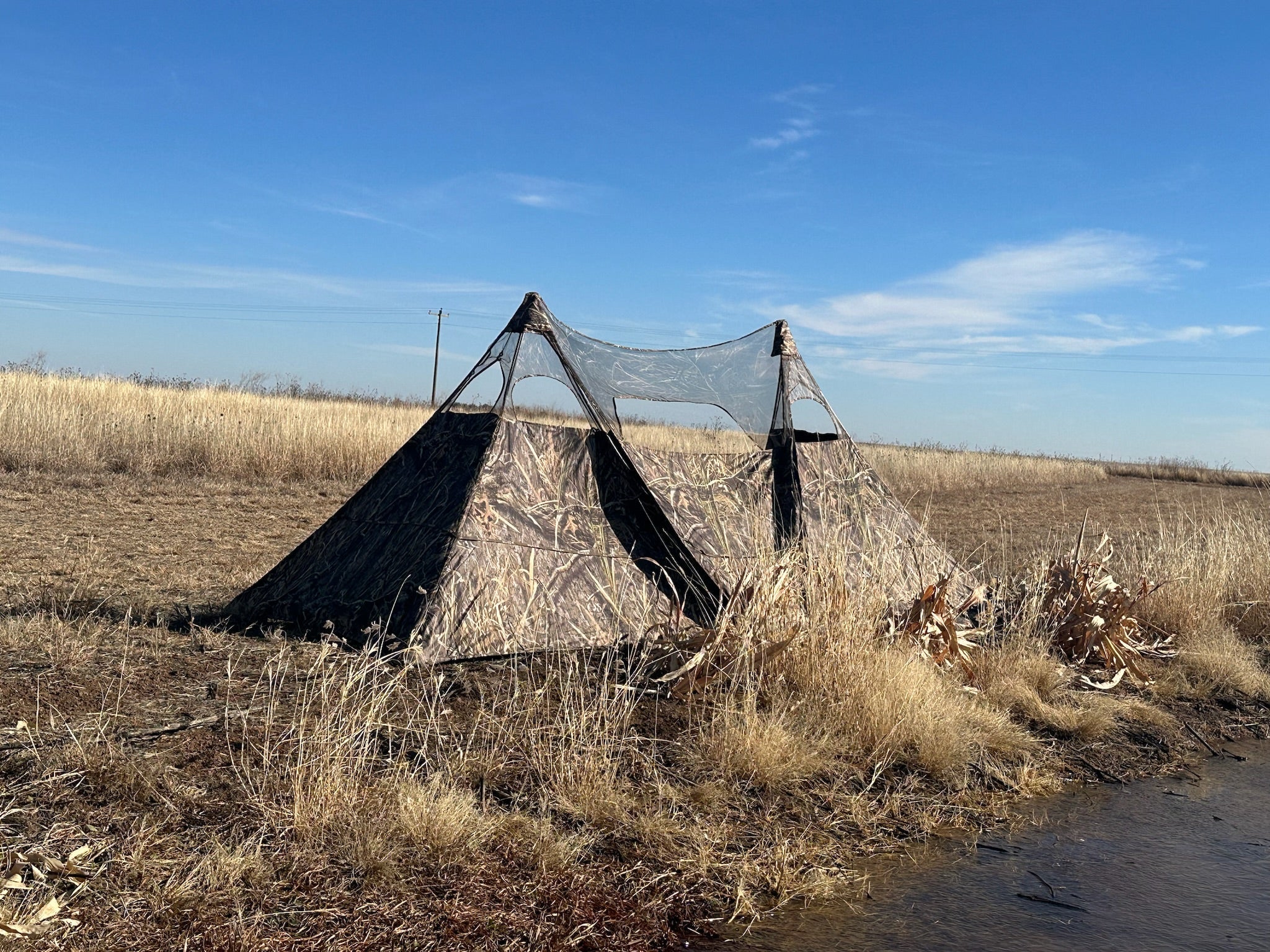 Camo hunting blind tent with mesh windows set up in dry field near water, ideal for waterfowl or deer hunting
