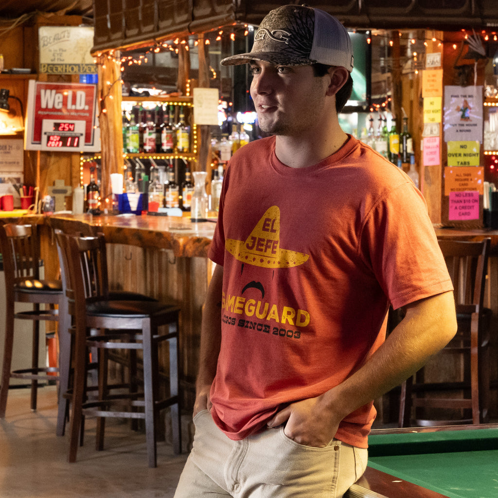 Man wearing orange El Jefe Gameguard t-shirt with sombrero graphic, camo trucker hat, in rustic bar setting