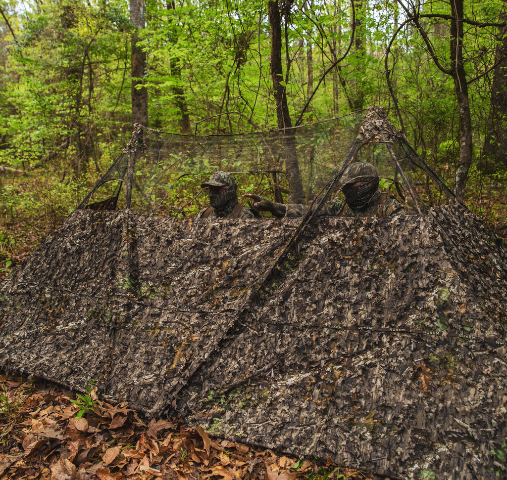 Two hunters in leafy camouflage blind with mesh windows, blending into forest for turkey or deer hunting.