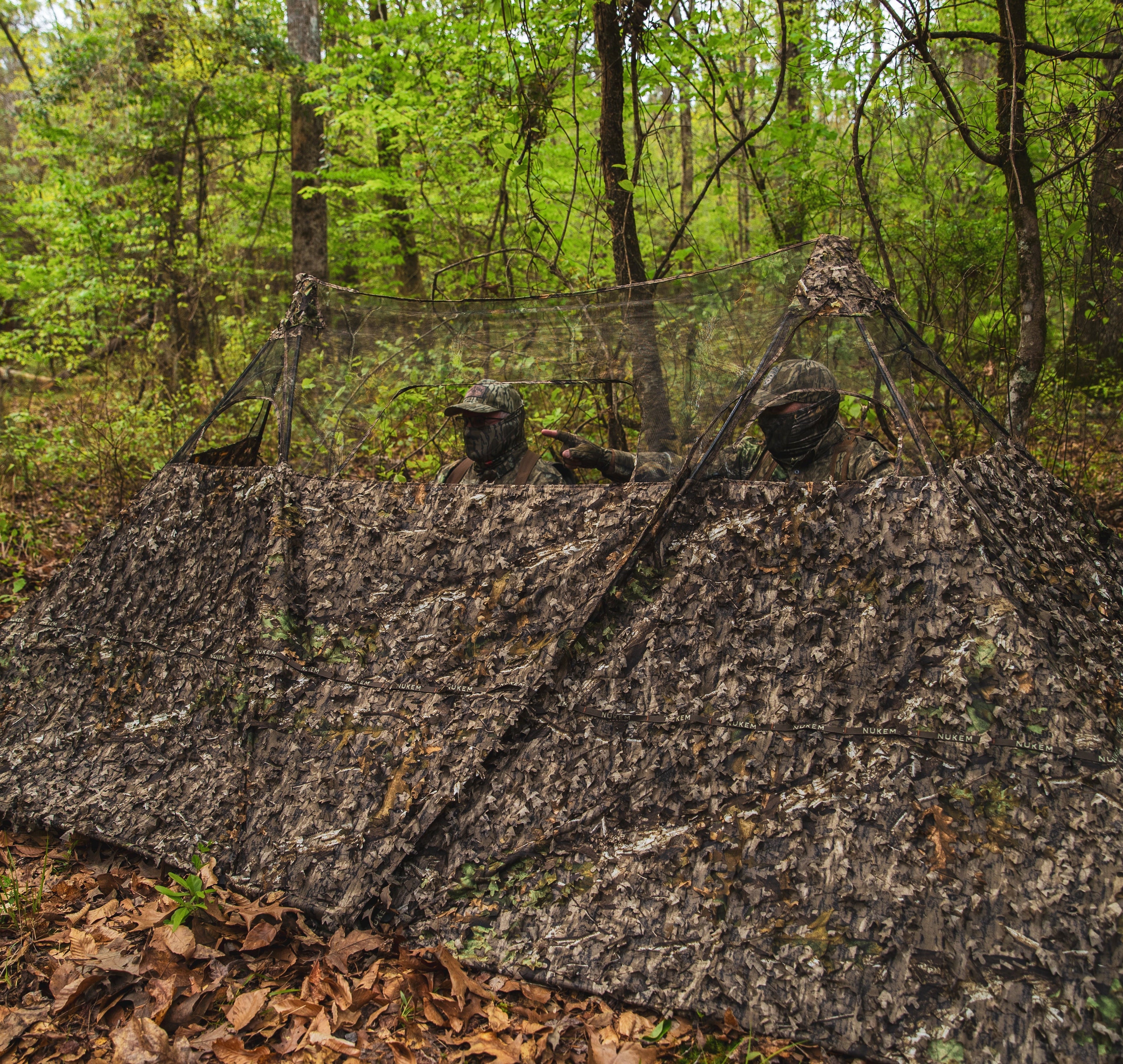 Two hunters in leafy camouflage blind with mesh windows, blending into forest for turkey or deer hunting.