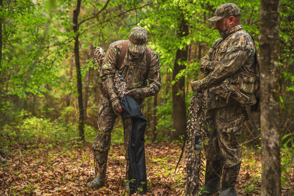 Two hunters in Mossy Oak camo gear prepare tree stand in forest, wearing waterproof pants and boots for outdoor hunting.