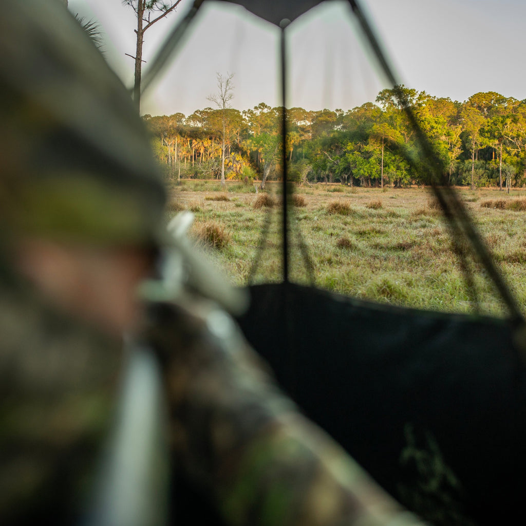 Hunter in camo gear aiming rifle from blind, overlooking grassy field and forest at dawn