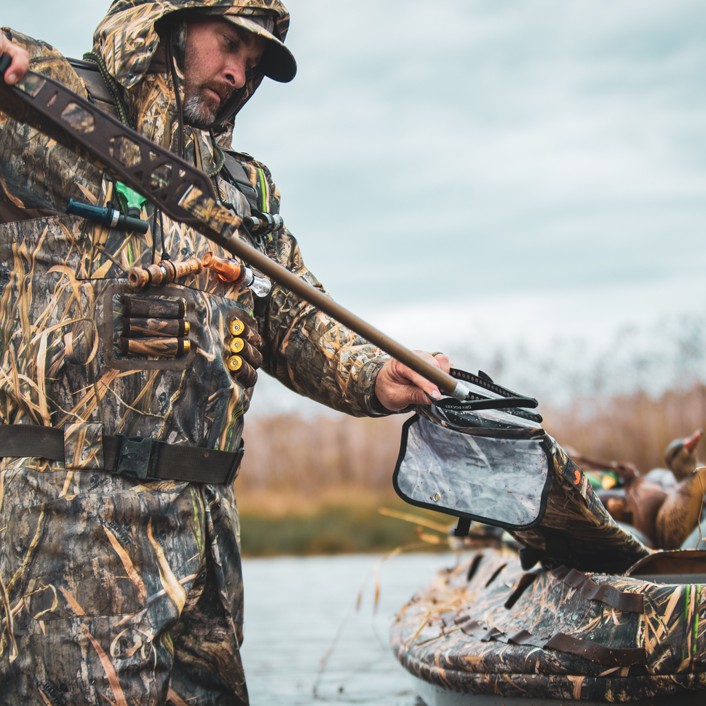 Hunter in Mossy Oak camo waders holding shotgun near duck blind on water, outdoor hunting gear