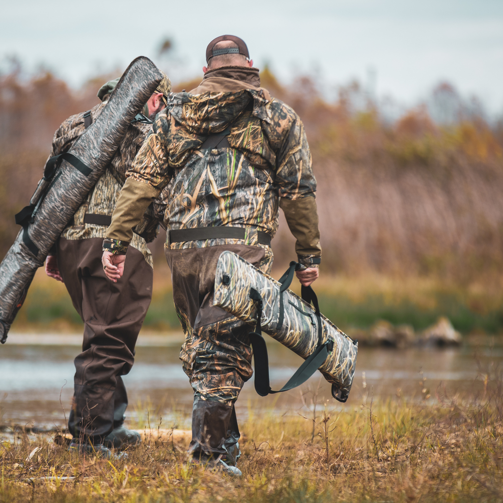 Two hunters in camo waders carrying shotgun cases through marshland, ideal for waterfowl hunting gear