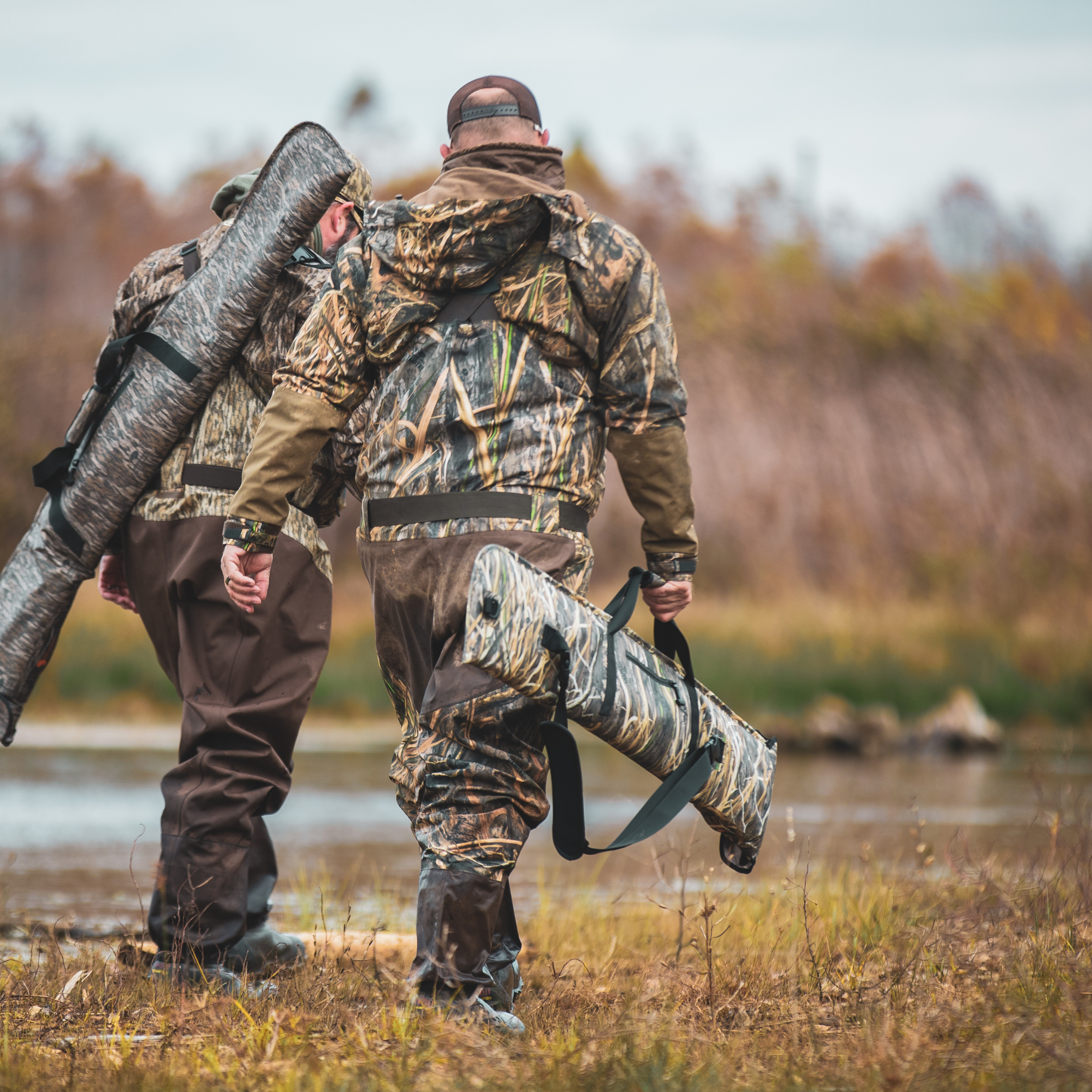 Two hunters in camo waders carrying shotgun cases through marshland, ideal for waterfowl hunting gear