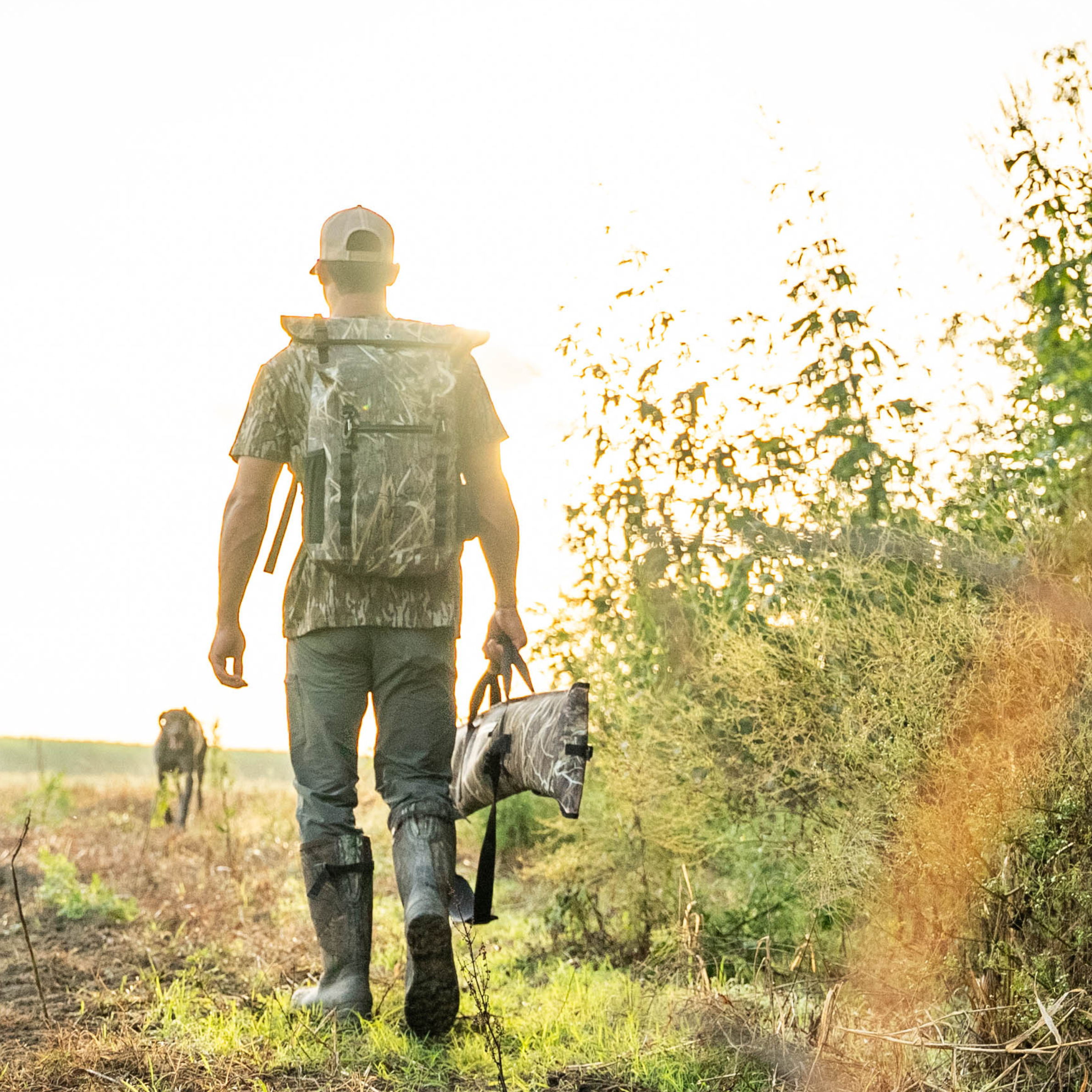 Hunter in camo shirt and boots carrying decoy through field at sunset