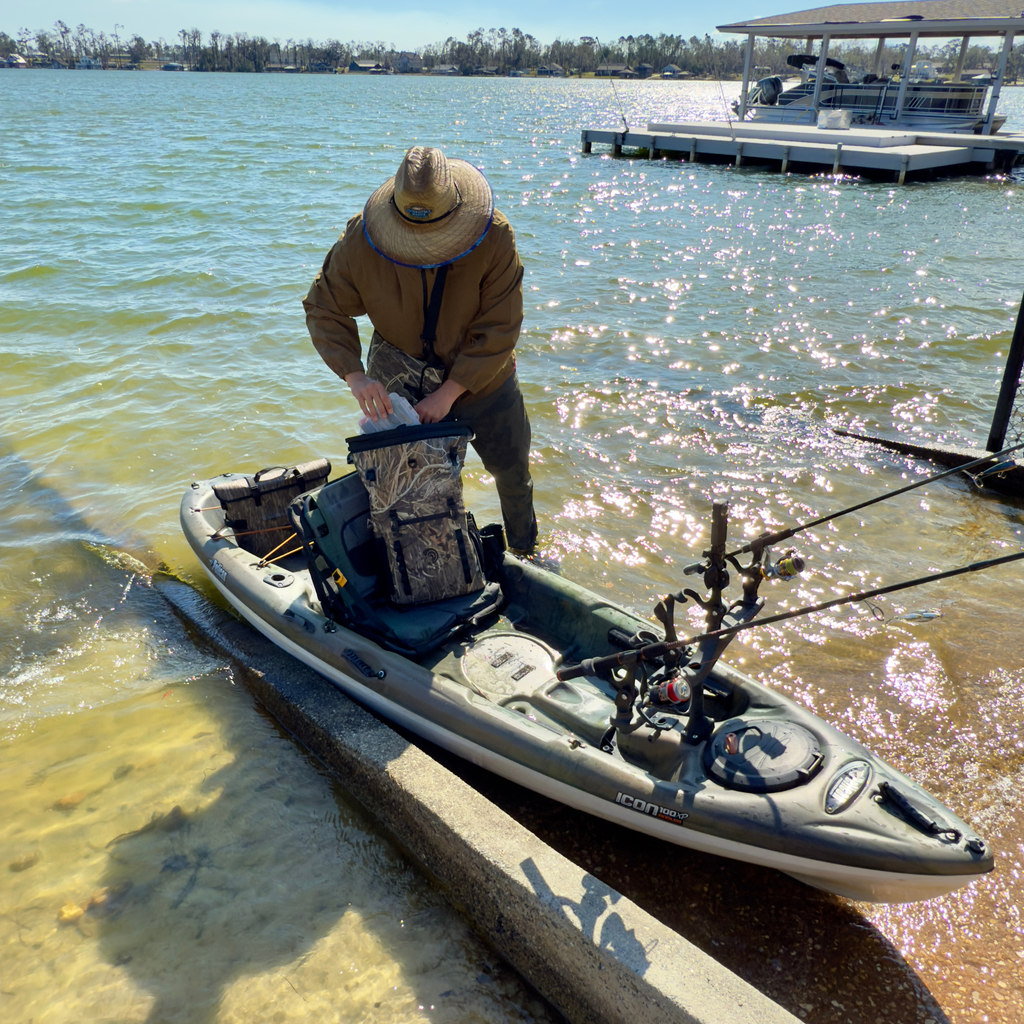 Fisherman in camo waders prepping fishing kayak with rod holders on sunny lake shore