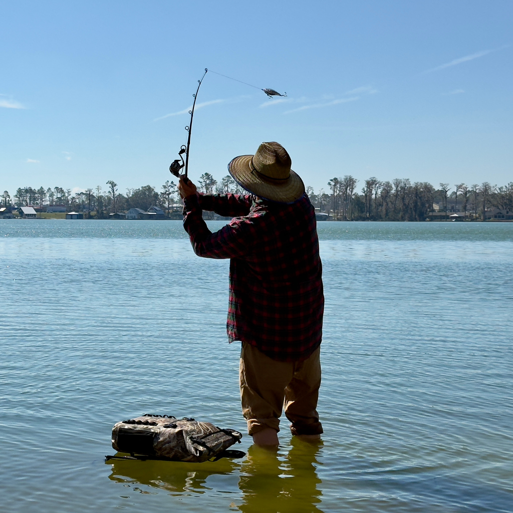 Angler casting fishing rod in shallow water, wearing plaid shirt, straw hat, and camo bag nearby