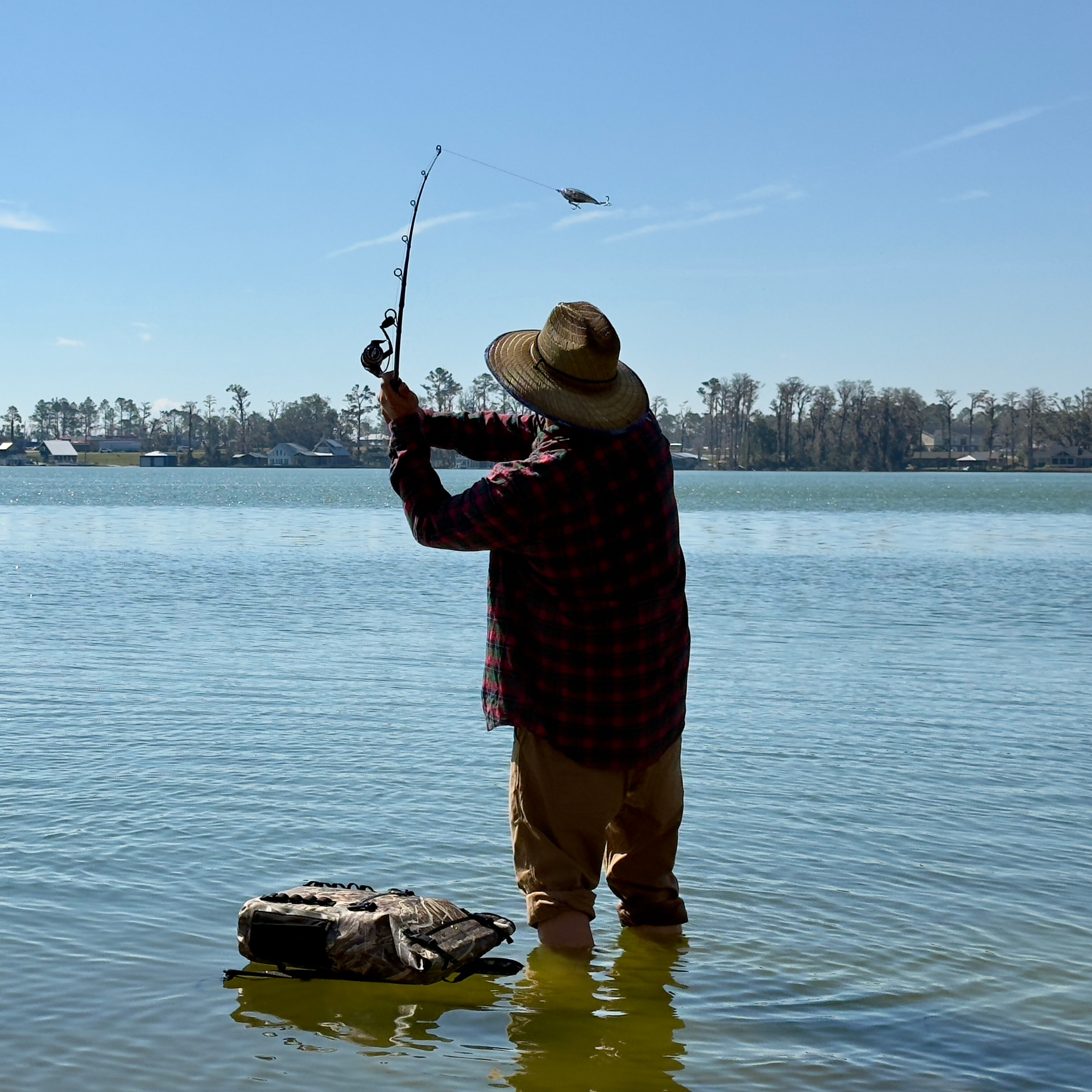 Angler casting fishing rod in shallow water, wearing plaid shirt, straw hat, and camo bag nearby