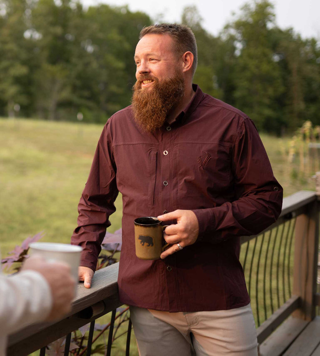 Man in maroon outdoor button-up shirt holding bear mug on deck, hunting apparel for men