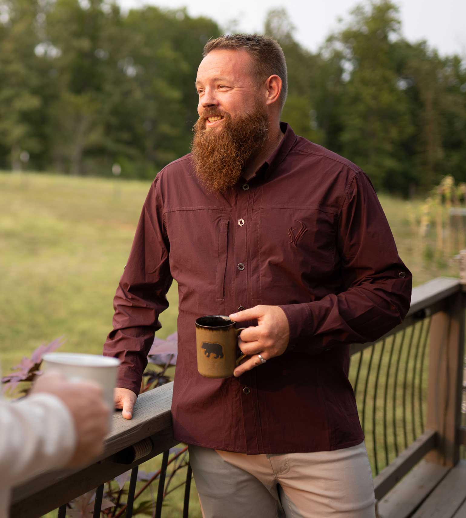 Man in maroon outdoor button-up shirt holding bear mug on deck, hunting apparel for men