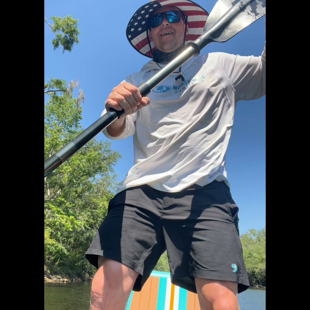 Man paddling kayak wearing American flag hat, Mossy Oak Fishing shirt, and black shorts on sunny river