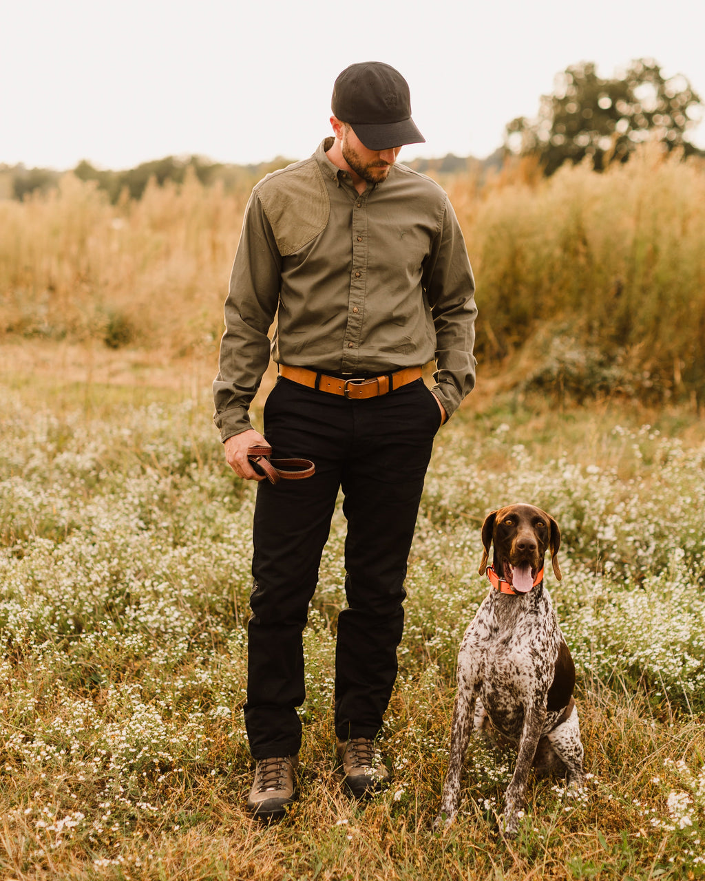 Man in the Ranger Green Jack Carr Hunters Shirt standing with a pointing dog in a meadow