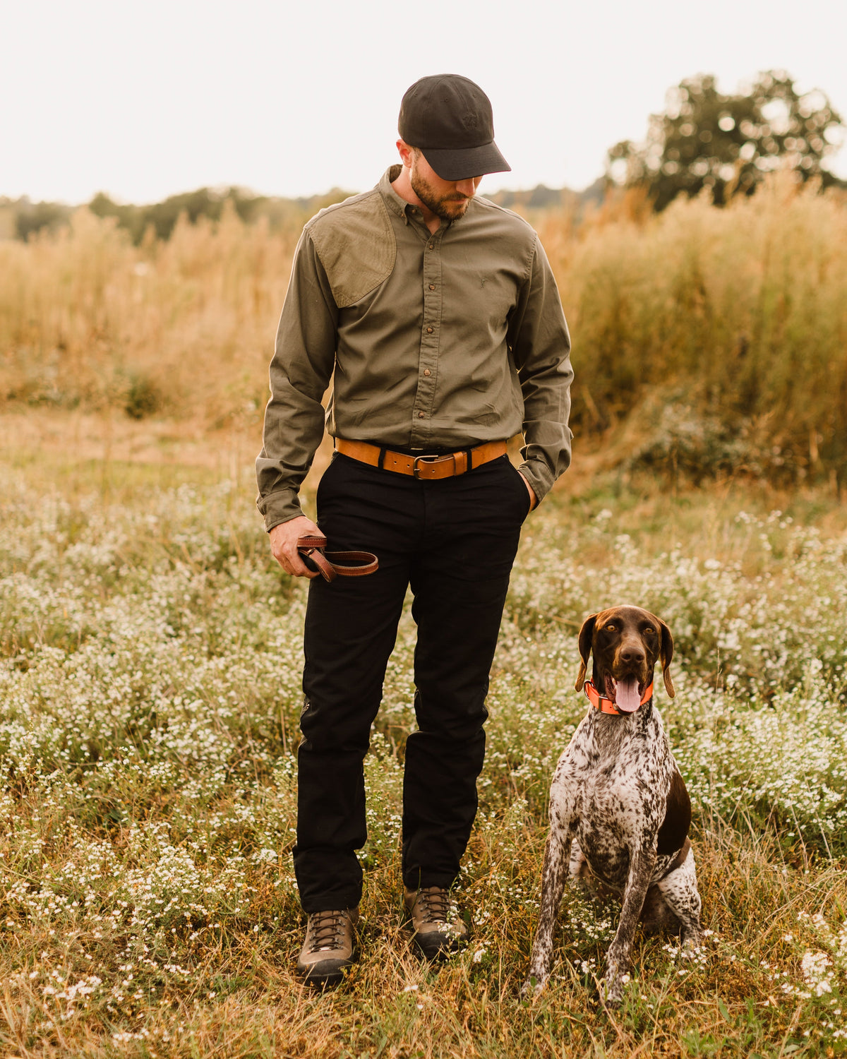Man in the Ranger Green Jack Carr Hunters Shirt standing with a pointing dog in a meadow