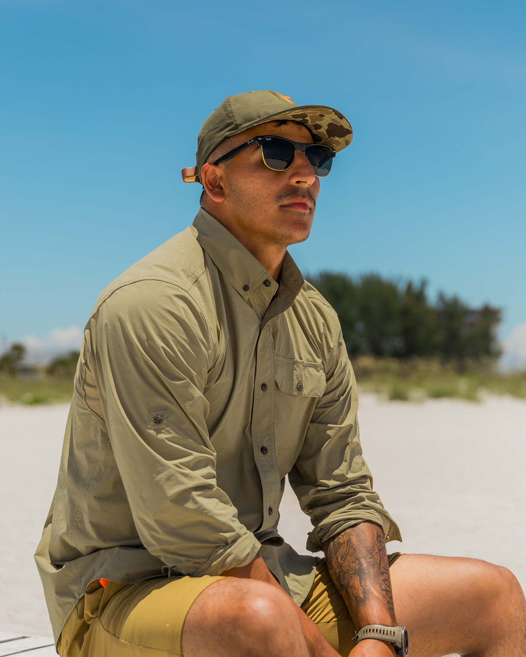 Man seated on a beach wearing the Tan Active+ Guide Shirt and sunglasses