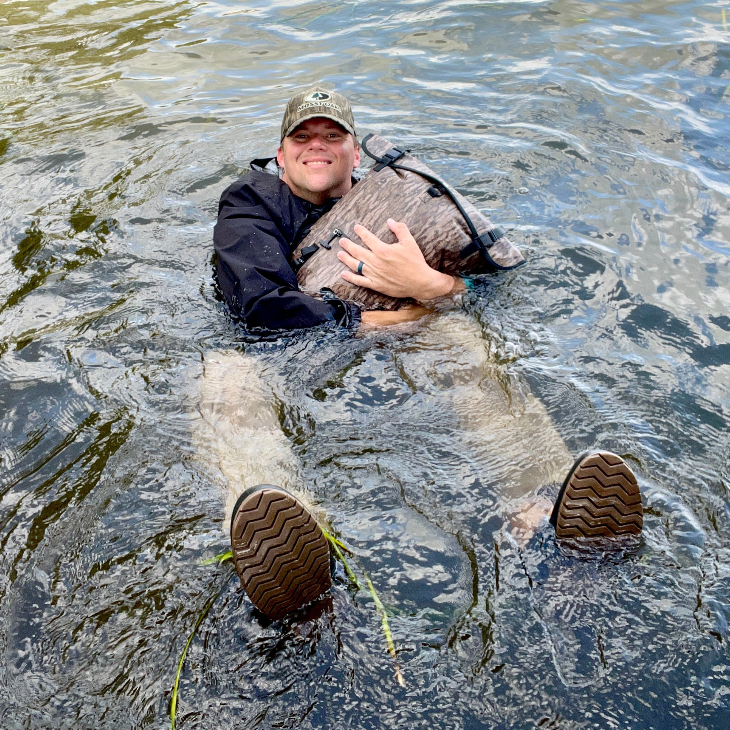 Man in water holding camo dry bag, wearing Mossy Oak hat and waterproof boots for hunting or fishing gear storage