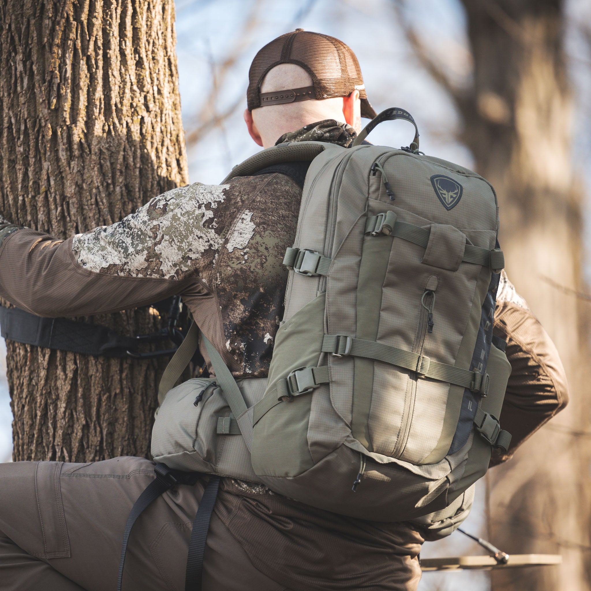Olive green hunting backpack with multiple straps and pockets, worn by hunter in camo gear near tree