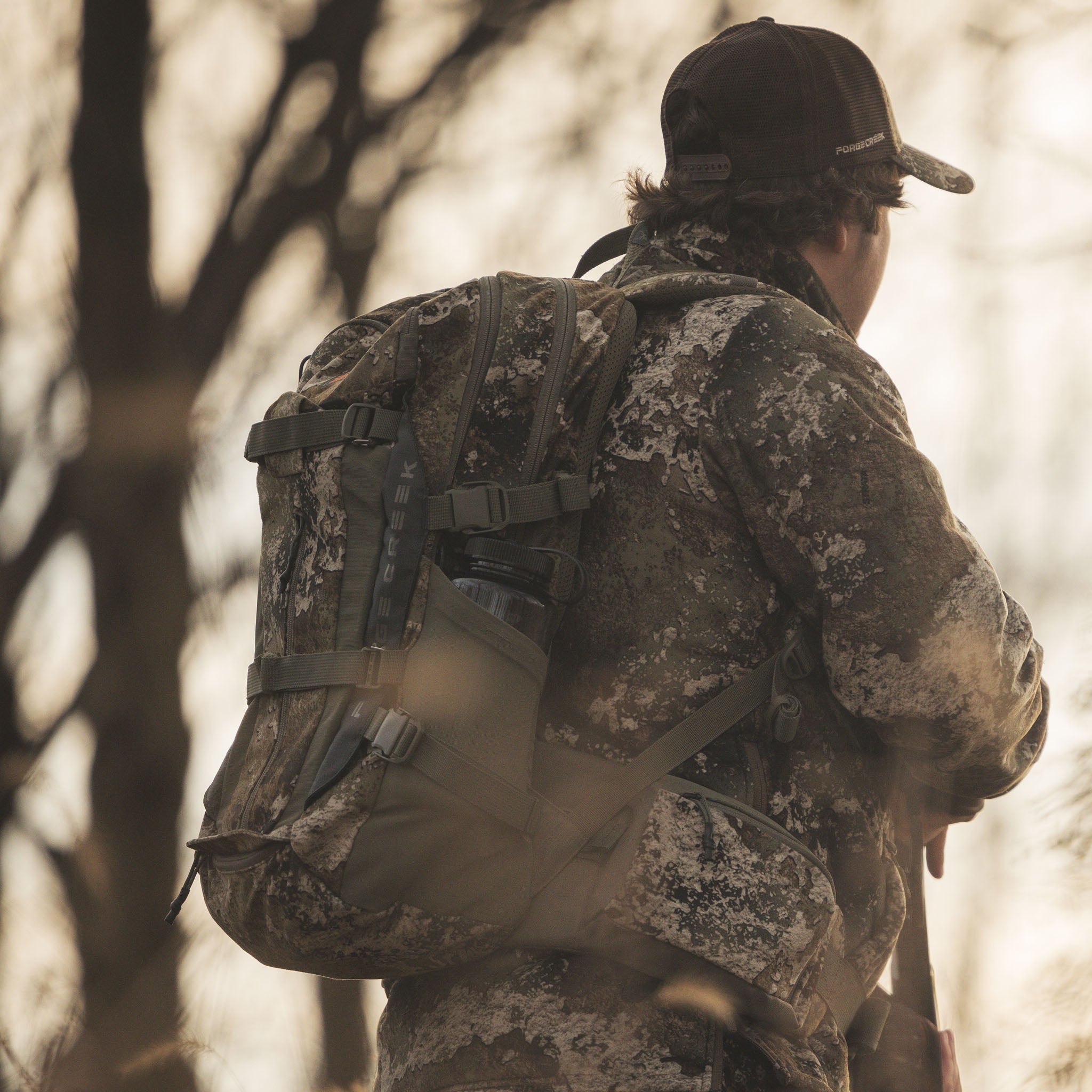 Hunter in camo jacket and backpack with water bottle, wearing ForgeCreek hat in woods
