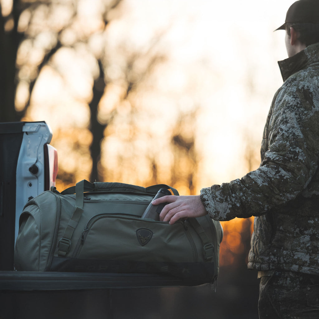 Hunter in camo gear packing olive duffel bag with binoculars at sunset, ready for outdoor adventure