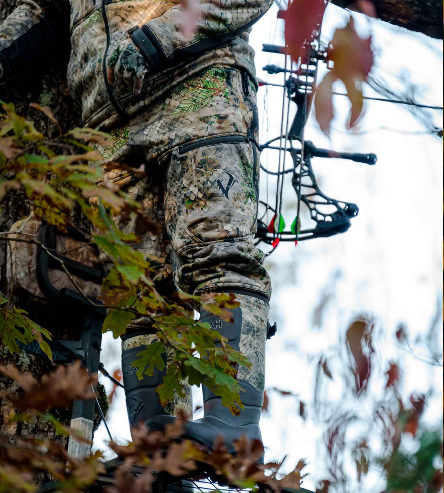 Bowhunter at full draw in a treestand wearing Vycah Kinsley Pants in Fall camo