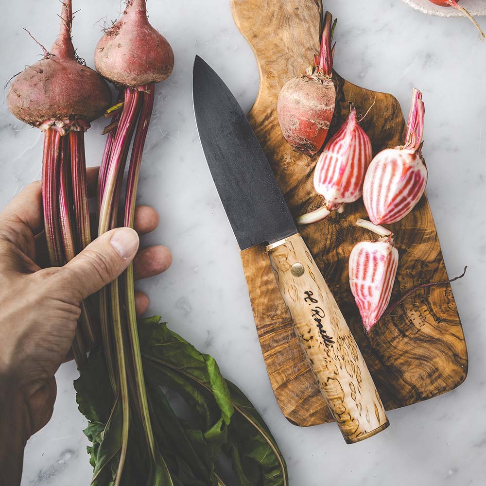 Hand holding fresh beets beside a chef knife with wooden handle on cutting board