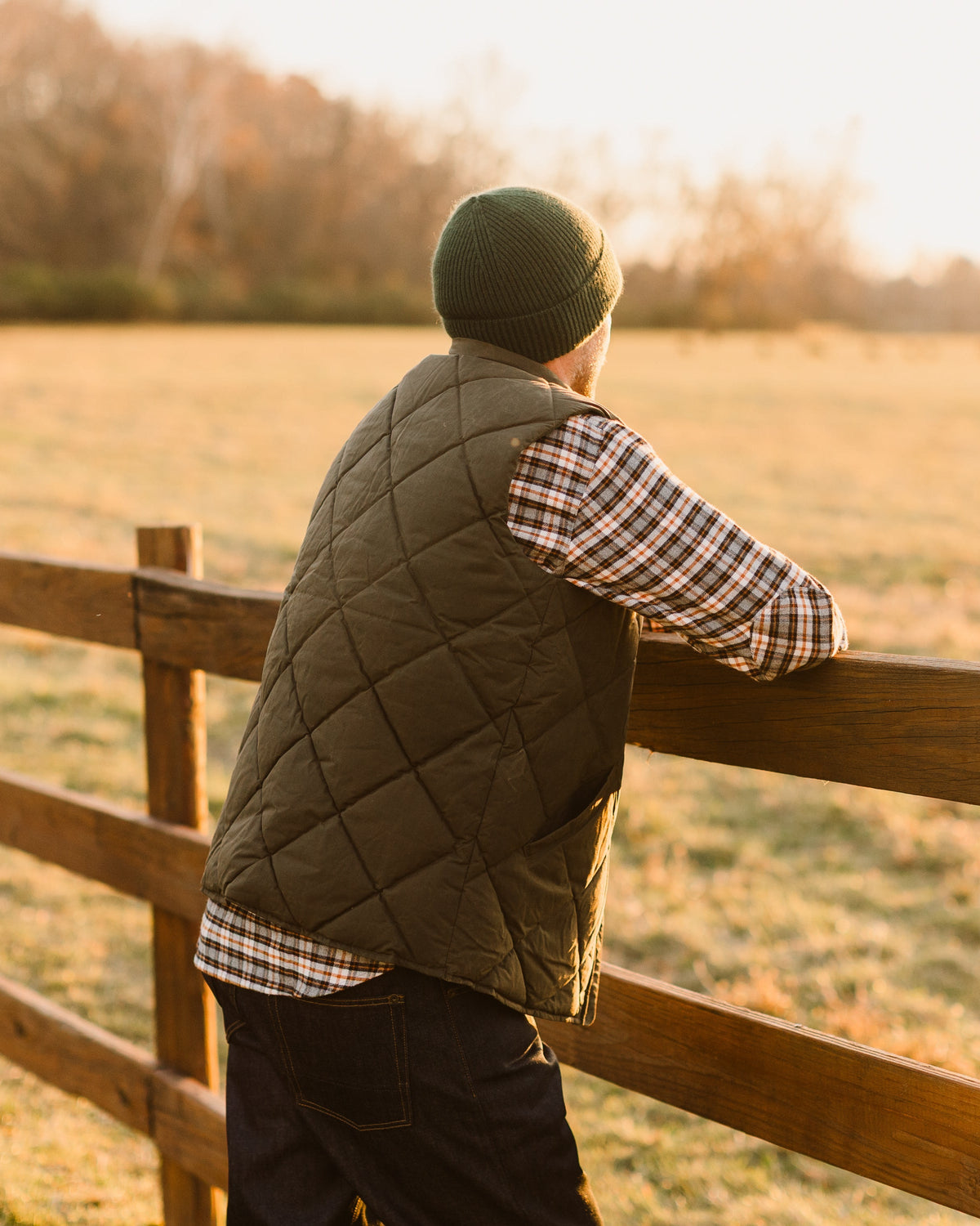 Loden Arthur Down Vest worn over plaid flannel, leaning on a split rail fence