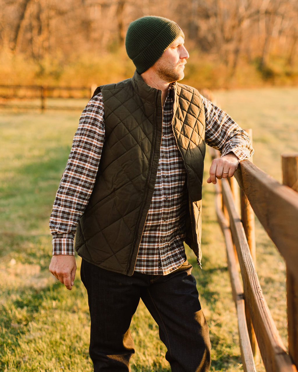 Man in a green beanie wearing the Loden Arthur Quilted Waxed Down Vest at a fence at sunrise