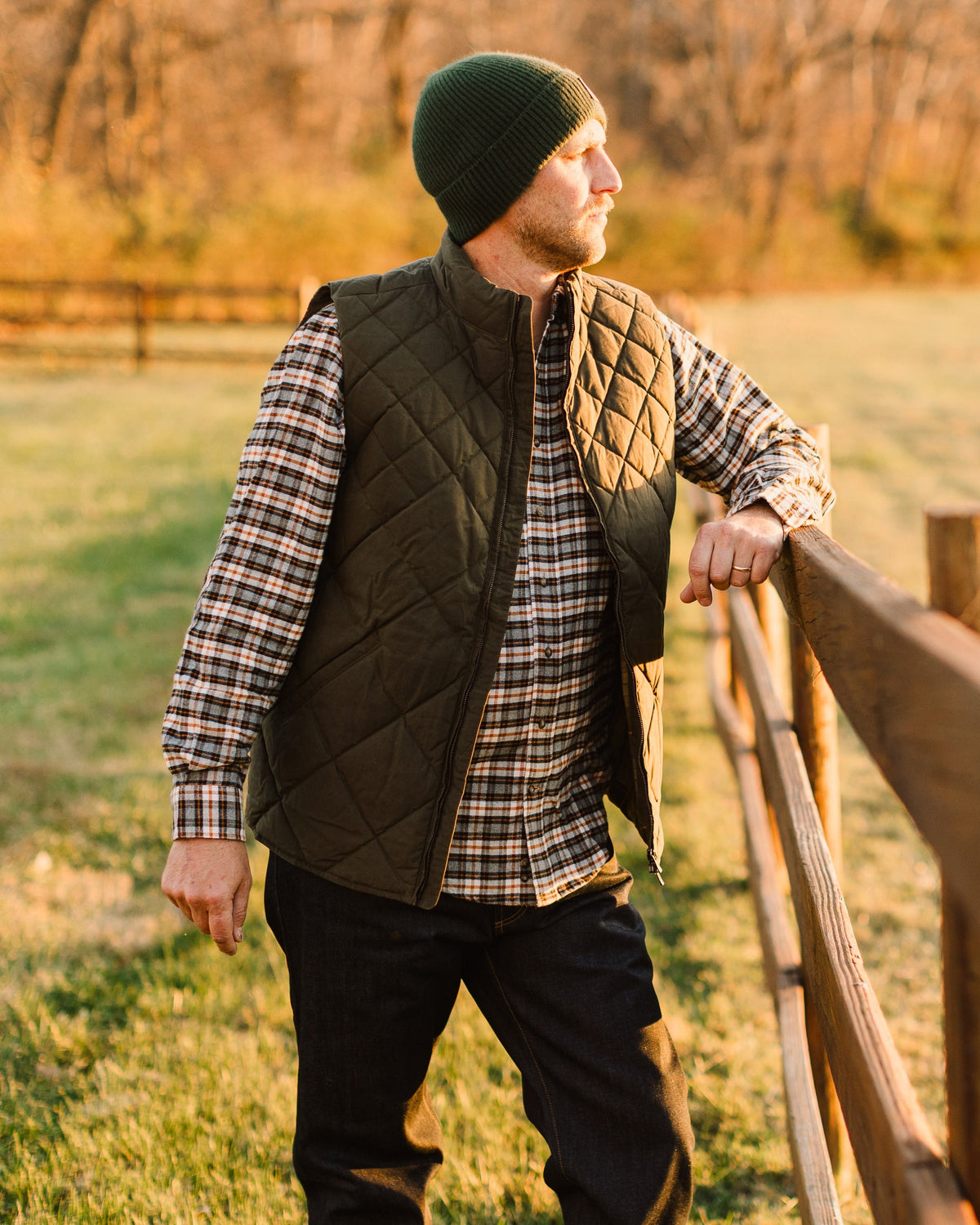 Man in a green beanie wearing the Loden Arthur Quilted Waxed Down Vest at a fence at sunrise
