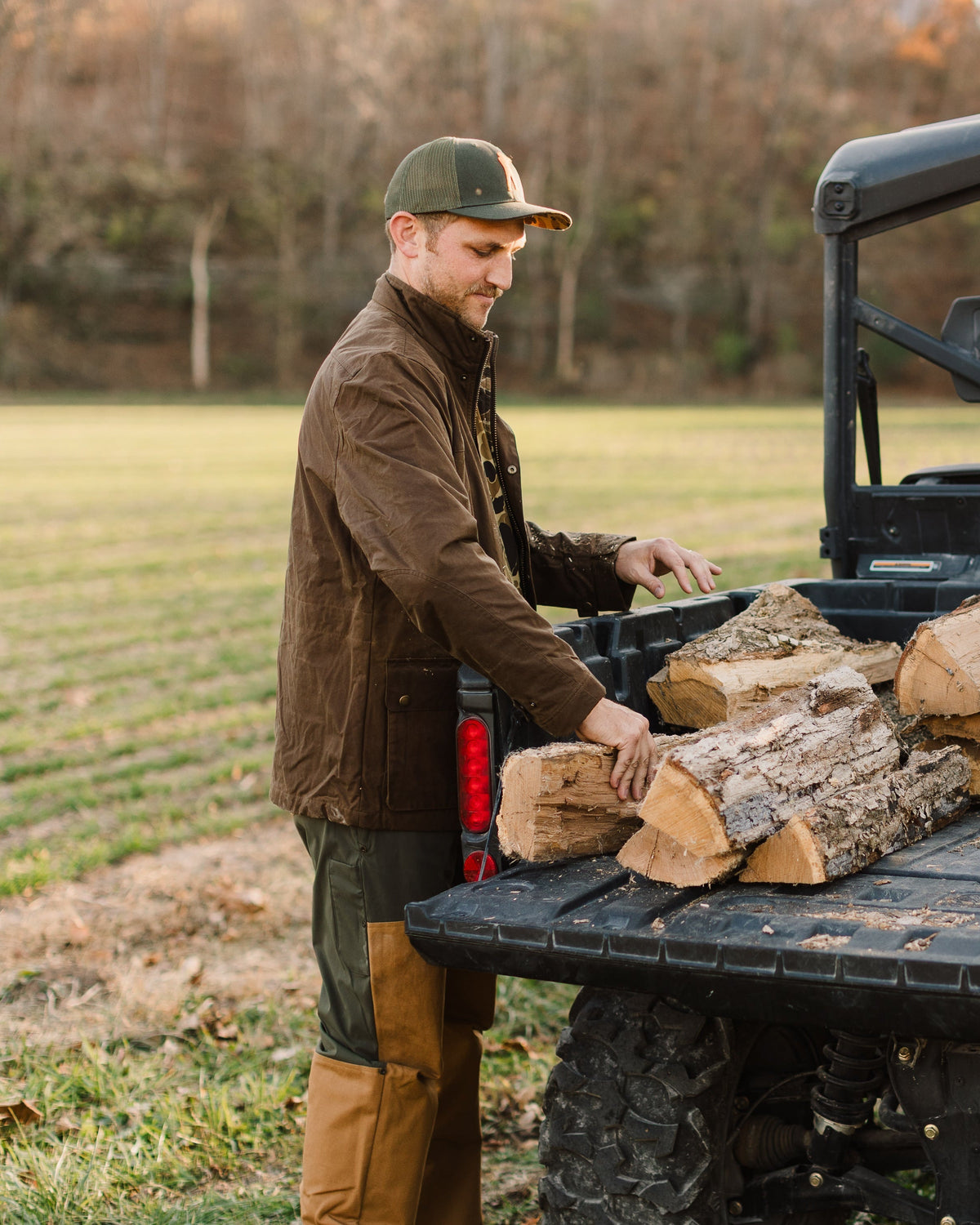 Man in the Oak Waxed Field Jacket and cap stacking firewood on a UTV in a field