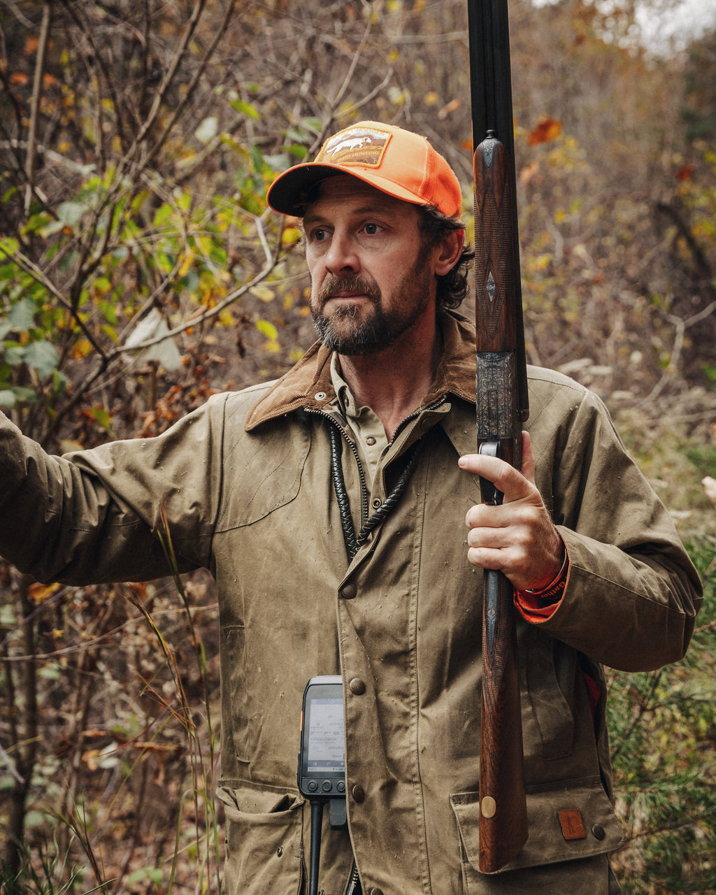 Hunter holding a side-by-side shotgun in the woods in the Olive Upland Jacket and blaze orange cap