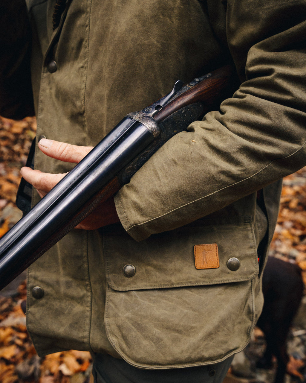 Hand pulling a shell from the pocket of the Olive Upland Jacket with a shotgun in hand