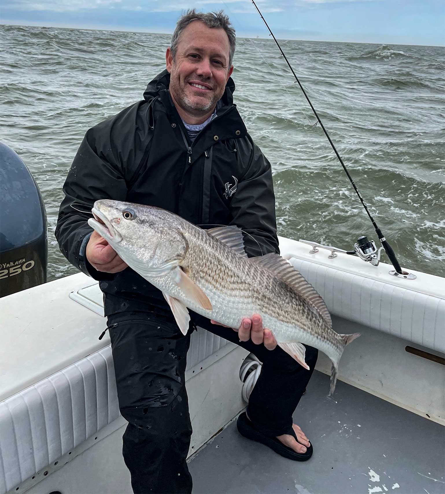 Man holding redfish on boat wearing black waterproof fishing jacket and pants, saltwater fishing gear